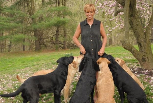 Woman standing outdoors surrounded by five attentive dogs.